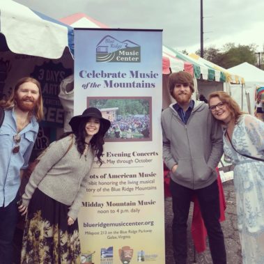 Four people smile in front of BRMC banner at Merlefest